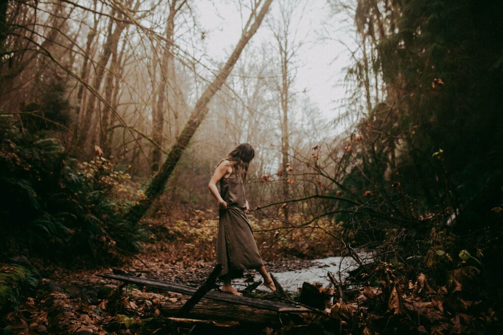 A woman uses a log to cross a stream in a foggy forest. She exemplifies several divine feminine archetypes.