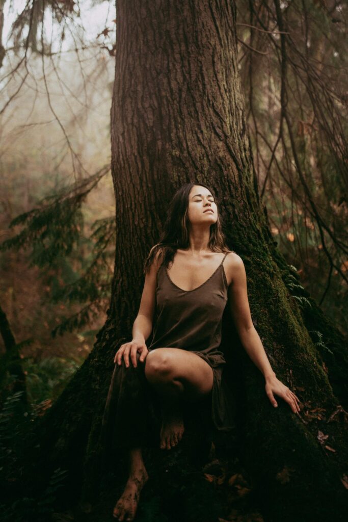 A woman contemplating the divine feminine archetypes during a quiet moment leaning against a tree.